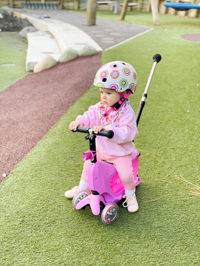 young toddler sitting on her pink mini2go deluxe ride on scooter with parents handle